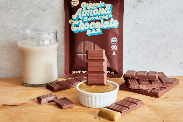 TJ's Organic Almond Beverage Chocolate Bar broken into pieces, dipped in sunflower seed butter, next to a glass of almond milk and product package in background